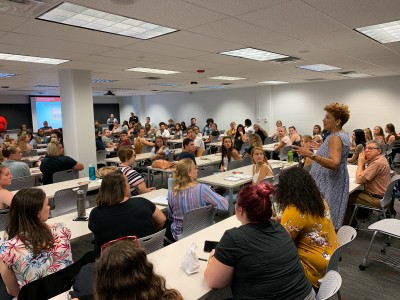 Students sitting at rows of tables look at a middle-aged Black woman who is standing and speaking to them.