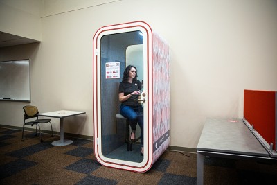 A woman wearing a mask and a shirt with an Ohio State Block O logo sits in a small, phonebooth-like pod.