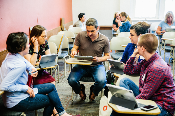 Instructors sit facing each other holding iPads and discussing course information