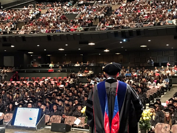 A man wearing academic regalia stands with his back to the camera in front of a crowd of people