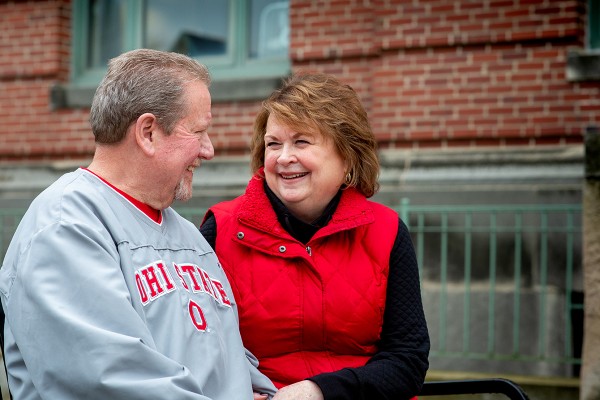 Senior couple sit on a bench outside, wearing red Ohio State gear and smiling at each other.