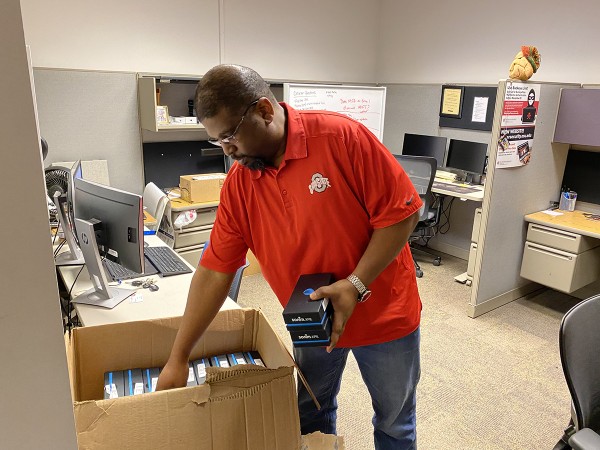 A man in a red shirt with the Ohio State logo stands in an office, looking through a cardboard box of wireless hotspots.
