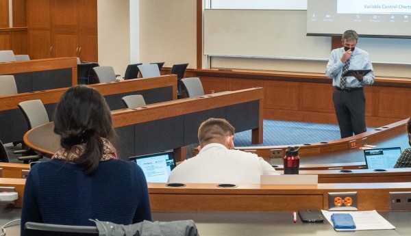 A professor wearing a mask stands in front of a few students in a classroom