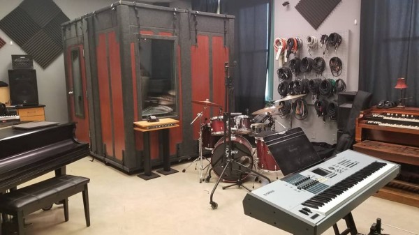A large red and black sound booth sits in the corner of a room filled with musical instruments, including a grand piano, a drum kit, an organ and a keyboard.