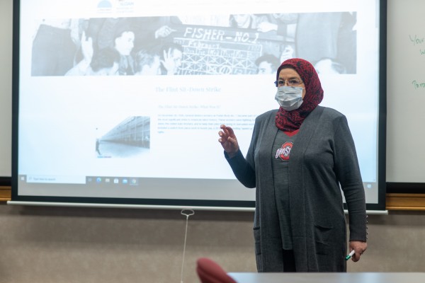 An instructor in a mask stands teaches her class with a presentation on the wall behind her