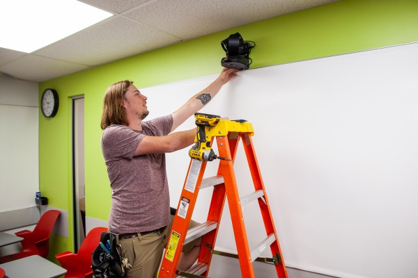 a staff member standing on a ladder adjusts a camera mounted on the wall of a classroom