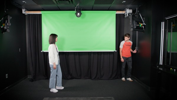 A woman wearing a white jacket watches as a young man lowers a green screen from the ceiling inside a video studio.