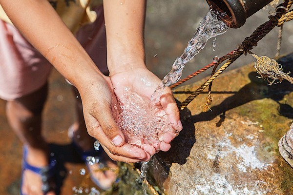 A young person cups their hands, filling them with clear water from a pump or fountain.