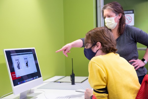 A student sits at a desk, working on a computer, while a woman standing behind him points over his shoulder to the screen