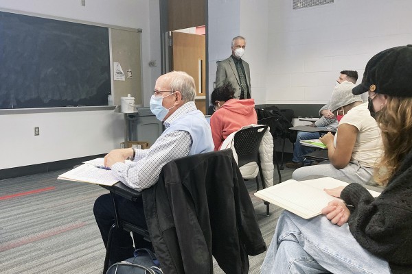 An older man with white hair sits in a desk at the front of a college classroom, wearing a surgical mask.