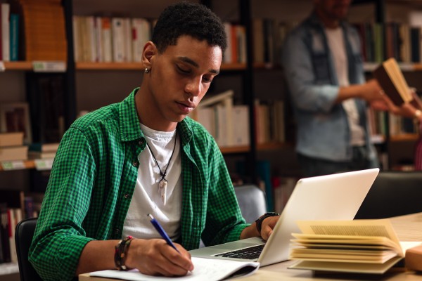 A student is in the library, working on an assignment using a laptop and a notebook.