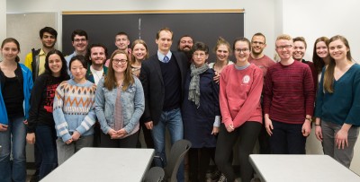 A professor stands with a group of students and an older woman with grey hair