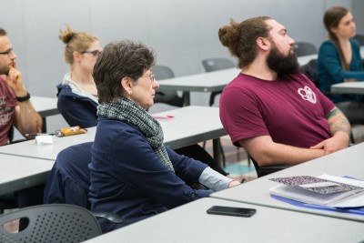 An older woman with grey hair sits at a table in a classroom next to a college student